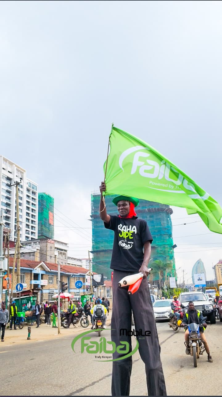 A performer on stilts waving a large green Faiba flag above the traffic at the Ngong Road junction