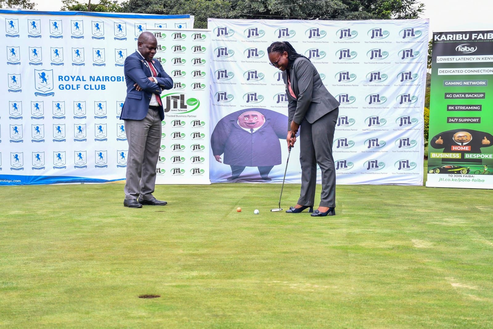 Two officials on the practice green at Royal Nairobi Golf Club, one putting in front of a JTL backdrop and a Karibu Faiba banner